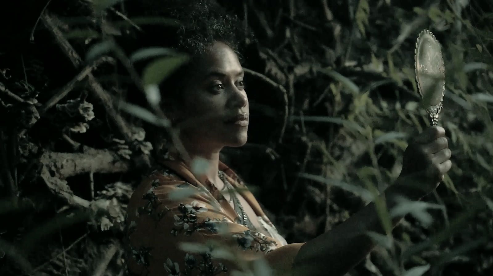 A still from the Neon Mirror video: a performer in dense foliage holding a hand mirror up to the light.