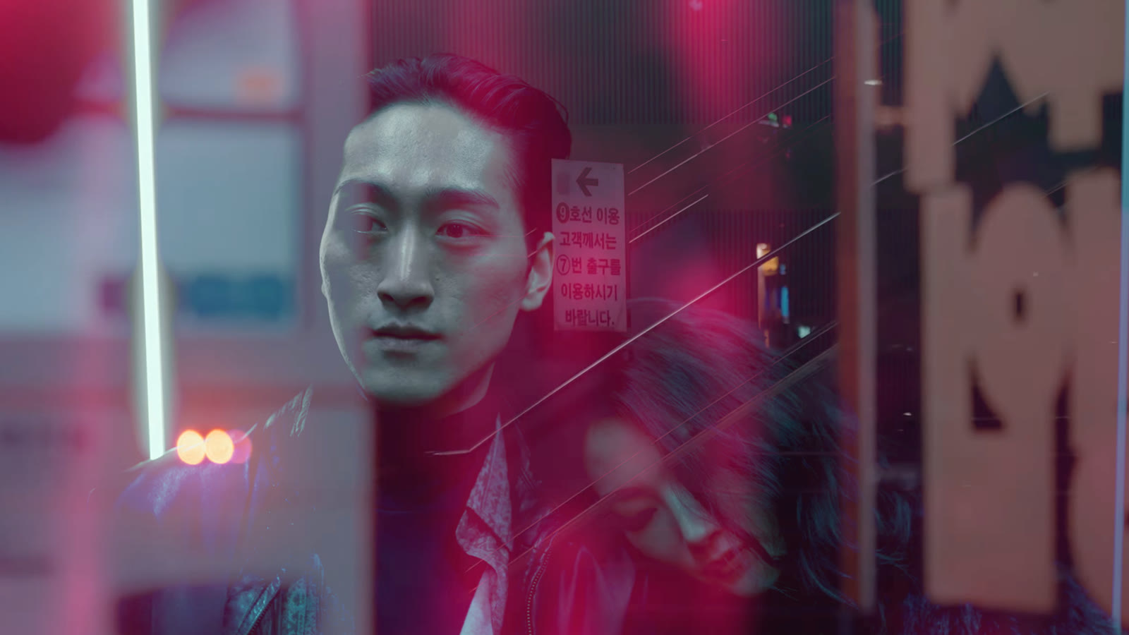 A still from the Starlit video: a performer lit red on a Seoul street at night, Korean subway signage and neon behind him, a second figure reflected in the glass.